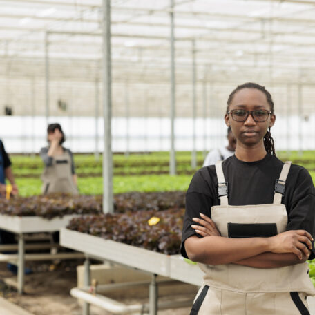 Portrait of confident african american greenhouse worker posing standing next to rows of organic vegetables seedlings. Professional woman standing in hydroponics microgreens plantation.